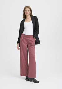 Kvinna som poserar i vit topp, svart blazer, dammig rosa vida byxor och svarta ankelboots mot en enkel vit bakgrund.