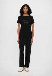 Black cotton t-shirt with short sleeves featuring a textured logo print. Paired with loose black trousers and light-colored sneakers.