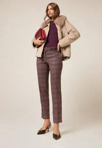 Manteau en laine beige avec col en fourrure, haut bordeaux, pantalon à carreaux dans des tons de rouge foncé et marron, et talons marron avec une boucle décorative.