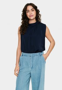 Top senza maniche blu scuro con dettagli plissettati sul collo, abbinato a pantaloni in denim azzurri a vita alta, che mostrano una vestibilità rilassata.