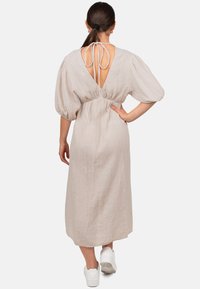 Beige, getirkt midi-jurk met pofmouwen en een diepe rugopening, cinched in de taille met een strikdetail. Draagt bij witte sneakers.