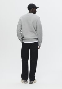 Gray sweatshirt with a crew neckline, ribbed cuffs, paired with black pants and beige sneakers. Model stands with back to the camera.