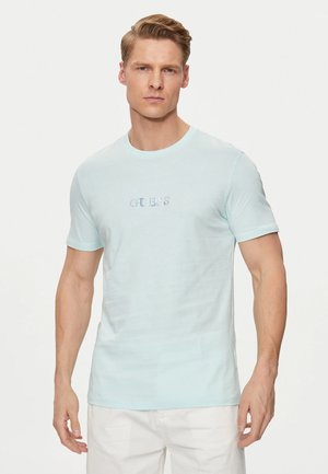 T-shirt en coton bleu clair à manches courtes, col rond, avec le logo "GUESS" brodé sur la poitrine. Texture lisse, coupe décontractée.