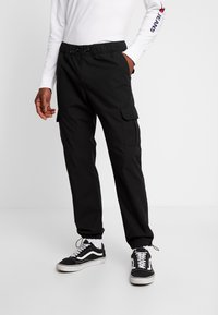 Pantaloni cargo negri cu talie elastică, buzunare laterale și manșete. Purtați cu adidași negri și un tricou alb cu mâneci lungi.