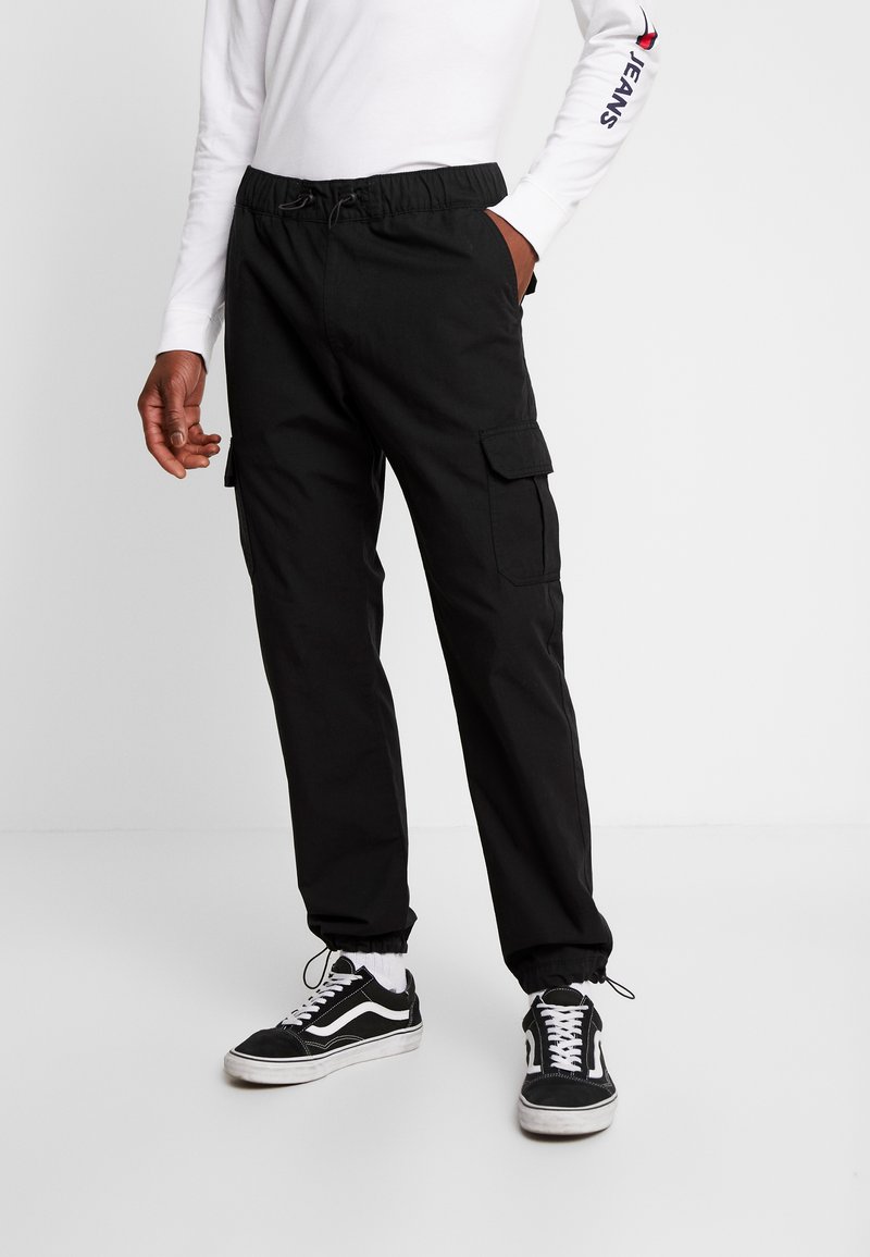 Pantaloni cargo negri cu talie elastică, buzunare laterale și manșete. Purtați cu adidași negri și un tricou alb cu mâneci lungi.