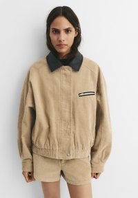 Corduroy-takki vaaleassa ruskeassa, mustalla nahkasella kauluksella, vetoketjullisella rintataskulla, elastisella vyötäröllä ja rentolla istuvuudella. Yhdistetty vastaavanvärisiin shortseihin.