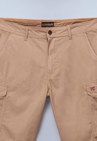 Bermudas de algodón beige con cierre de botón, dos bolsillos laterales y un pequeño parche de bandera. La cinturilla presenta costuras naranjas.