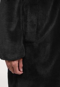 Zwarte, pluche stof met een zachte textuur; het beeld toont een hand die een donker kledingstuk vasthoudt met een zijzakdetail en een manchetkraag.