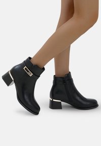 Botas de tobillo de cuero negro con tacón cuadrado, que presentan un hebilla y detalles en tono dorado, cierre de cremallera y un diseño de puntera redondeada.