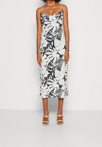 Robe imprimée de feuilles tropicales en noir et blanc, sans manches avec de fines bretelles, longueur mi-mollet, texture du tissu lisse. Portée avec des sandales à talons nude.