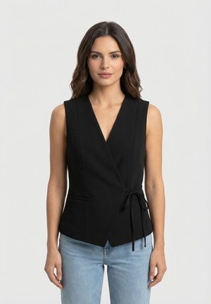 Mujer con cabello castaño ondulado, vestida con una blusa negra sin mangas con lazo lateral y jeans azul claro, de pie contra un fondo claro y sencillo.