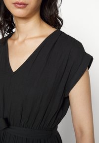 Femme portant une robe noire à encolure en V, manches courtes et ceinture à la taille, montrant le buste et le cou sur un fond blanc.