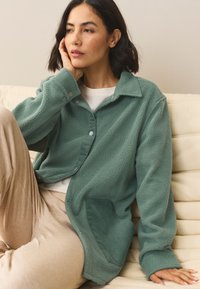 Camicia in pile con chiusura a bottoni, di colore verde petrolio tenue, con bottoni a pressione, tasca in velluto a coste e vestibilità ampia, abbinata a pantaloni beige.