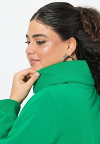 Felpa verde con colletto alto, dotata di una texture morbida e polsini a costine. Modello mostrato con una mano sul colletto, indossando grandi orecchini a cerchio in argento.
