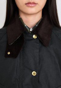 Black jacket with a corduroy brown collar. Gold snap buttons secure the front, layered over a green plaid shirt.