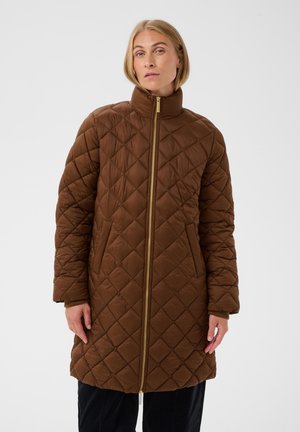 Femme portant un manteau matelassé marron longueur genoux avec un col montant et une fermeture éclair devant, debout devant un fond clair uni.