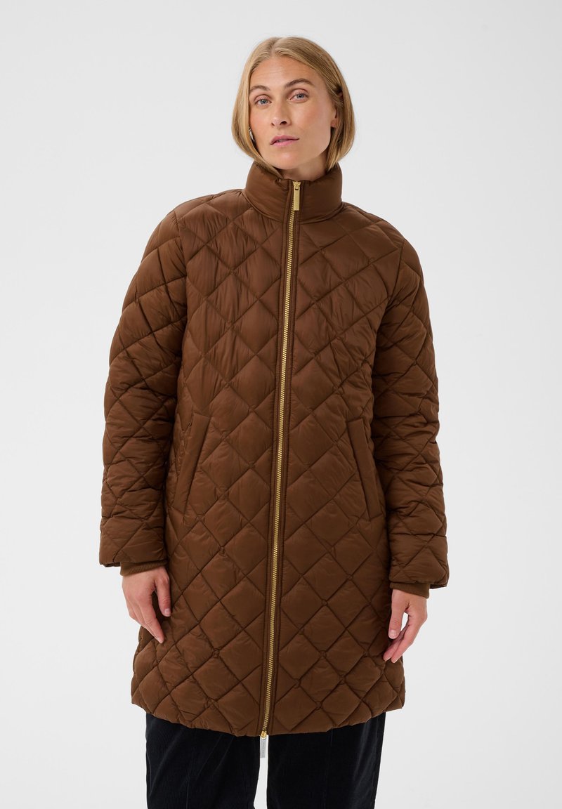 Femme portant un manteau matelassé marron longueur genoux avec un col montant et une fermeture éclair devant, debout devant un fond clair uni.