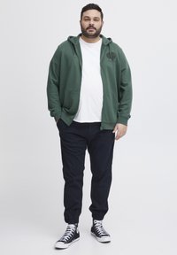Felpa verde con cappuccio e tasche frontali sopra una maglietta bianca, abbinata a pantaloni scuri e sneaker alte nere.