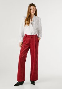 Sarkanas korduroja platās kājas bikses ar piegriezumiem, valkātas kopā ar baltu garām piedurknēm blūzi ar volānu apkakli. Melni augstpapēžu potītes zābaki.