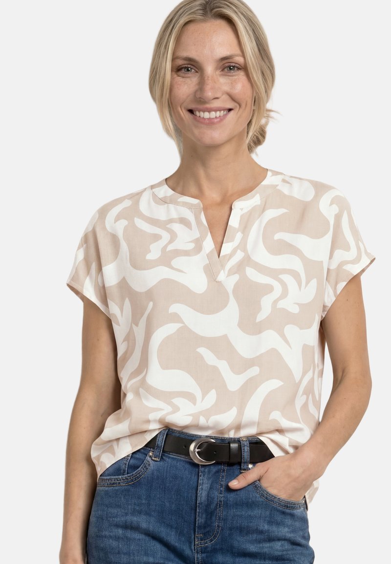 Mujer sonriente con cabello rubio lleva una blusa beige y blanca de manga corta con estampado, vaqueros azules y un cinturón negro con hebilla plateada.