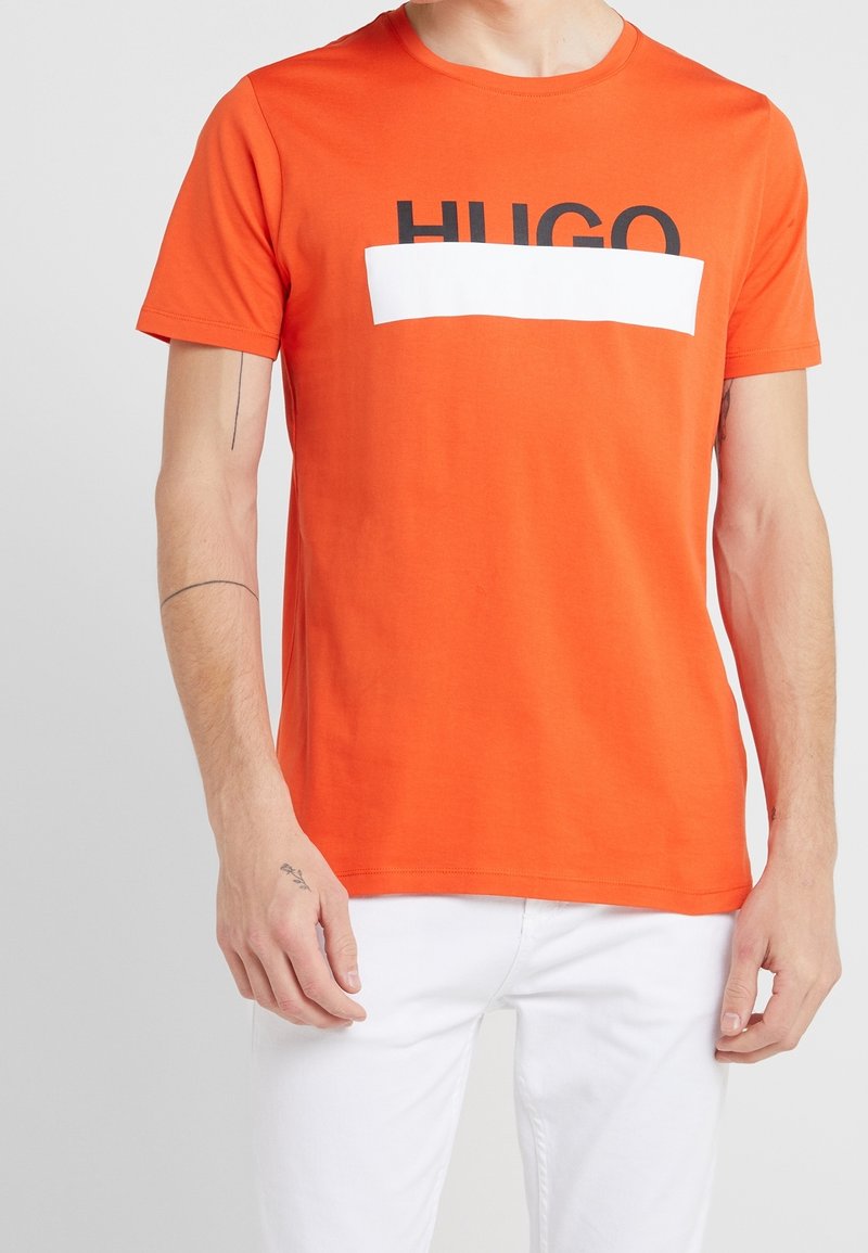 T-shirt en coton orange avec le texte "HUGO" en noir et un accent rectangulaire blanc traversant la poitrine. Col rond et manches courtes.