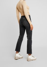 Jeans de mezclilla negros con corte recto y ligero desgaste, combinados con zapatillas texturizadas blancas. La parte superior es una camiseta ajustada de manga larga en color beige.