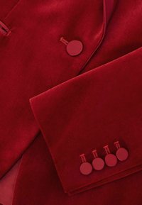 Rød fløjl blazer med en glat tekstur. Har en rund knaplukning og fire dekorative knapper på ærmet.