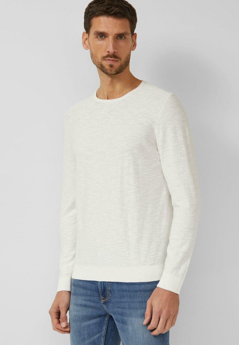 Man die een witte textuursweater met lange mouwen en een blauwe spijkerbroek draagt, staand tegen een effen lichtgrijze achtergrond.
