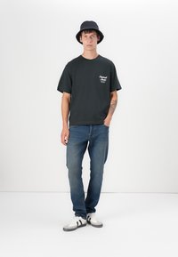 Black cotton t-shirt with a front logo, paired with blue denim jeans and black bucket hat. White sneakers feature black stripes.