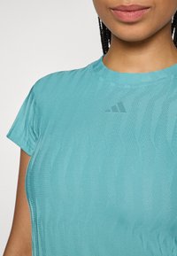 Tyrkysové sportovní tričko s krátkým rukávem, s texturovaným vzorem a malým šedým logem na levé straně hrudi.