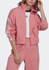 Veste zippée rose en tissu doux avec trois rayures blanches sur les manches, poignets côtelés et pantalons assortis, conçue pour un usage décontracté.