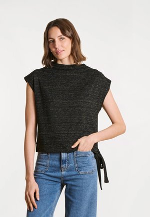 Femme aux cheveux bruns mi-longs portant un haut noir sans manches avec un nœud sur le côté et un jean bleu, posant avec une main dans la poche.