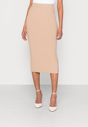 Kvinde iført en beige ribstrikket midi-nederdel og hvide spidse højhælede sko med ankelremme, stående mod en ensfarvet baggrund.