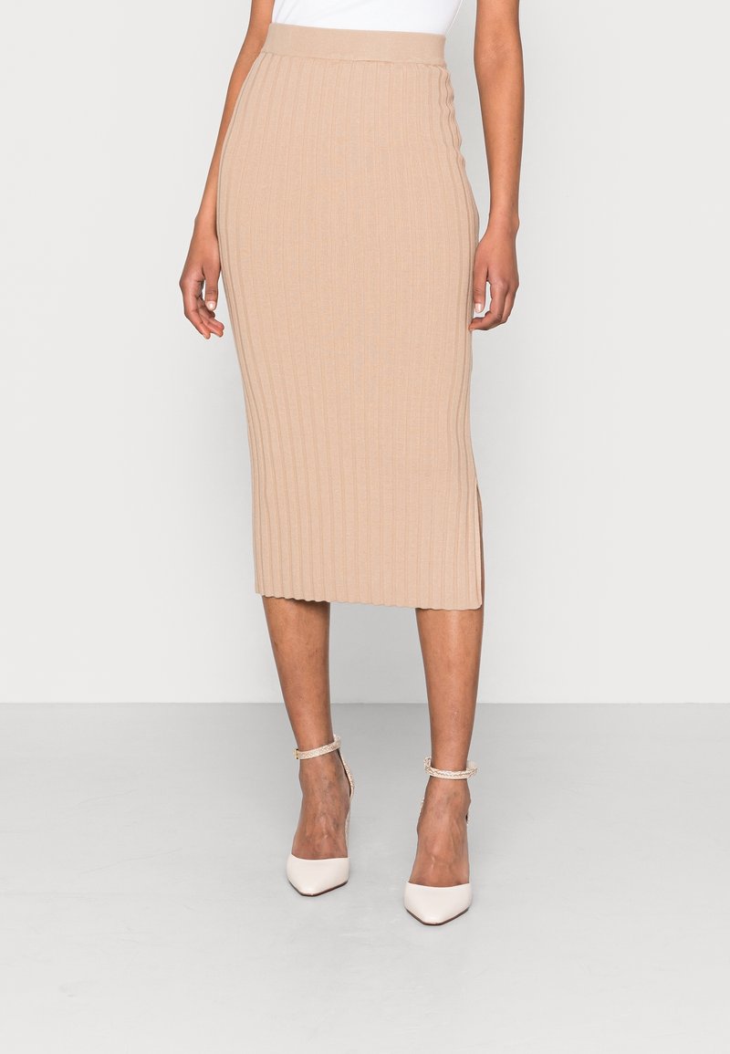 Femme portant une jupe midi beige en maille côtelée et des talons hauts blancs pointus avec des brides à la cheville, debout devant un fond uni.