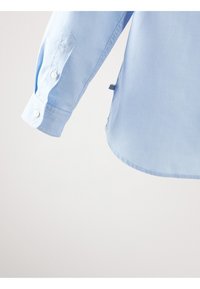 Lichtblauwe lange mouwen shirtmanchet met witte knopen en een klein zwart-wit gestreept stoffen label aan de zijnaad.