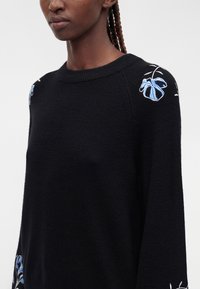 Pull en maille noire avec broderie florale bleue et blanche sur les épaules et le haut des manches, porté par une personne aux cheveux tressés.
