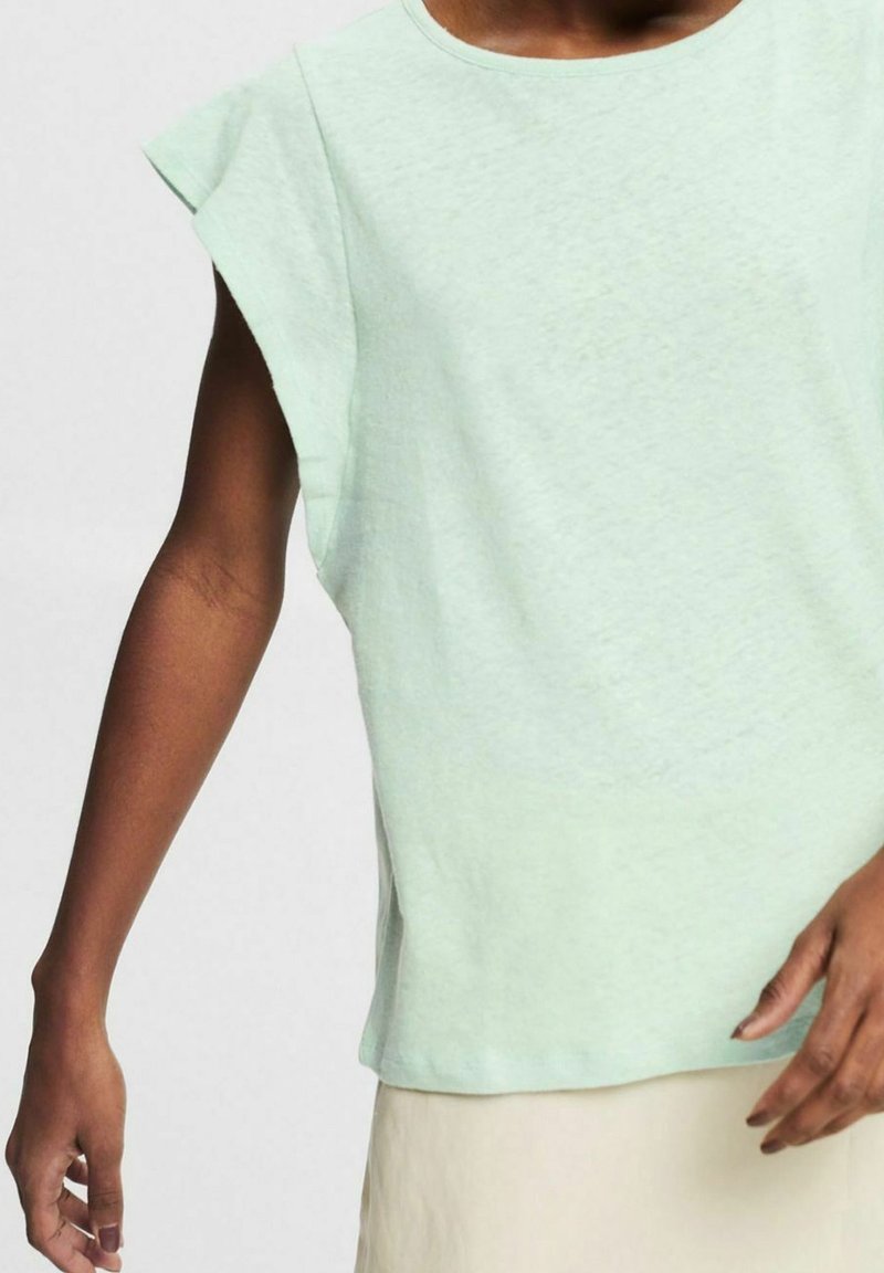 Personne portant un haut sans manches vert clair et un bas beige, les bras détendus le long du corps sur un fond uni.