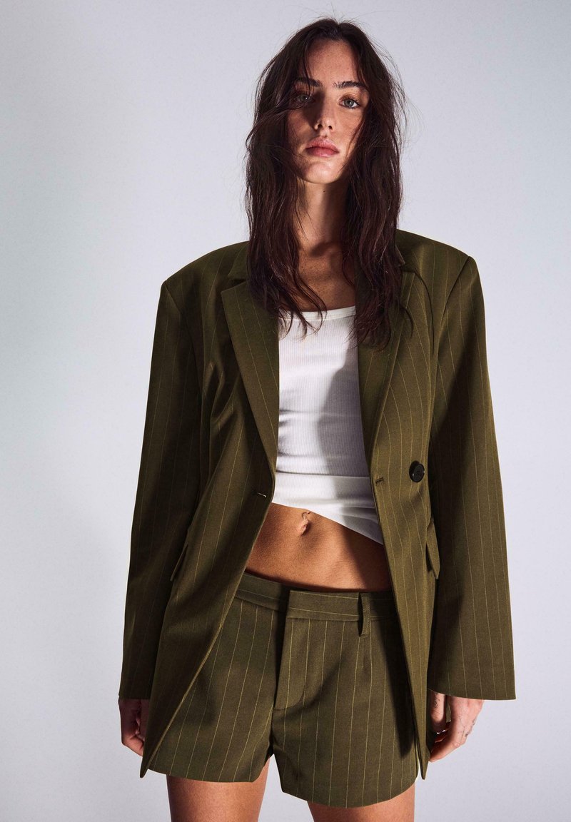 Femme aux longs cheveux noirs portant un blazer oversize à rayures vert olive et un short assorti, sur un débardeur blanc court.