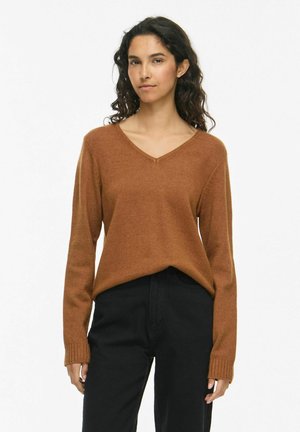 Femme aux cheveux bouclés foncés portant un pull marron à col en V et un pantalon noir, debout devant un fond blanc uni.