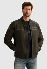 Mann mit kurzem braunem Haar und Bart, der eine dunkelgrüne Lederjacke mit Reißverschluss und Abzeichen sowie hellblaue Jeans trägt, die Hände in den Taschen.