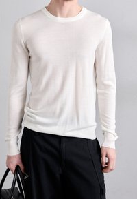 Leichter, cremefarbener Strickpullover mit geripptem Hals- und Ärmelbund. Figurbetontes Design mit langen Ärmeln und glatter Textur. Kombiniert mit dunklen Hosen.