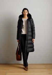 Vrouw in een lange zwarte pufferjas, crème trui, zwarte broek en bruine laarzen, met een bruine suède handtas.