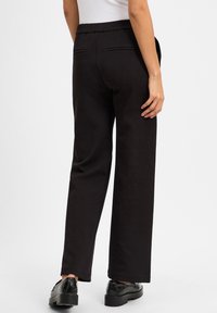 Zwarte, wijde broek met een elastische tailleband, twee achterzakken en een gladde textuur. Onder de enkel lengte, gecombineerd met glanzende zwarte schoenen.