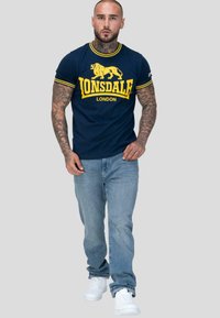 Muž s tetováním oblečený v námořnickém modrém tričku Lonsdale London, světle modrých džínách a bílých teniskách, kráčející vpřed na šedém pozadí.