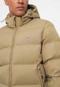 Beige puffatakki, jossa on vetoketju ja säädettävä huppu. Rinnassa logo ja sivussa vetoketjulliset taskut. Kangas on sileä ja pehmustettu.