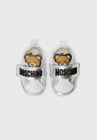 Silverfärgade barnskor med en slät metallisk yta, försedd med ett svart spänne med texten "MOSCHINO" och en björnmotivit som kikar fram från insidan.