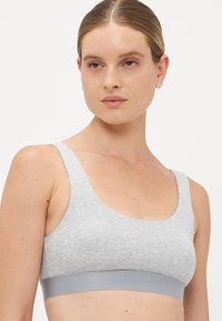 Young woman wearing a light grey sports bra with wide straps and a solid grey elastic band, posing against a plain white background.