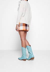 Mujer vestida con un suéter blanco, falda a cuadros en naranja y marrón, botas azules de estilo vaquero hasta la mitad de la pantorrilla, y un anillo azul en la mano izquierda, de pie de lado.