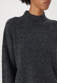 Maglione grigio scuro lavorato a maglia con un colletto alto e a coste e una vestibilità ampia. Il tessuto ha una consistenza morbida e un motivo sottile in tutto il capo.