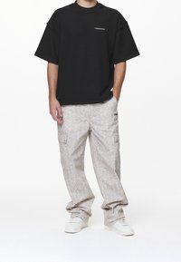 Man in een zwart oversized t-shirt en losse lichtbeige cargo broek met witte sneakers, met één hand in zijn zak.